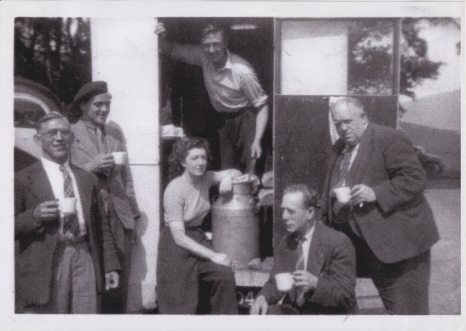 A film unit gathered around a milk churn outside a catering vehicle, drinking tea from cups and saucers